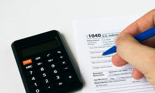 A closeup of a person's hand filling up a 1040 tax form next to a calculator.