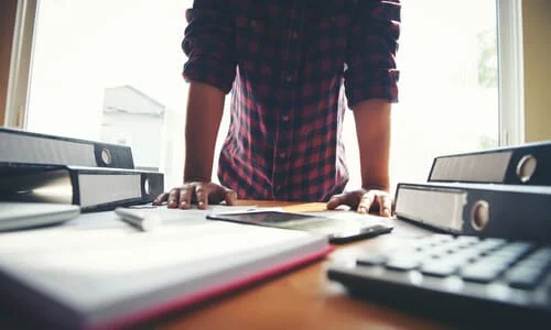 A person in his study with both hands on his desk having trouble dealing with tax records scattered around him.