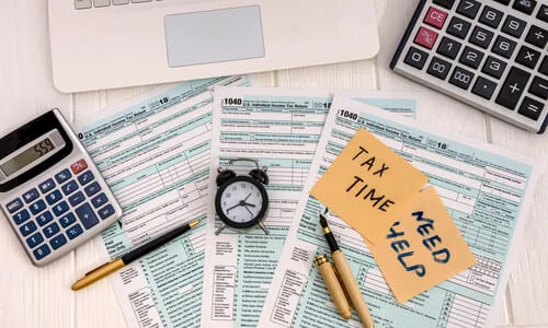 Tax forms and calculators scattered on a table with sticky notes and an alarm clock.