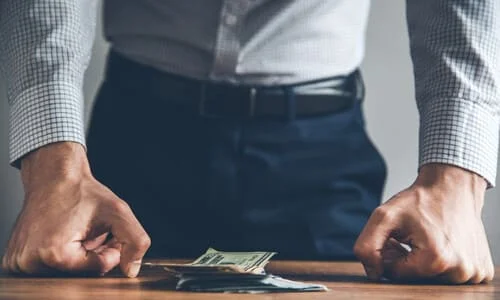 A man worried about his finances holding his fists against a table and looking down at a small stack of money.