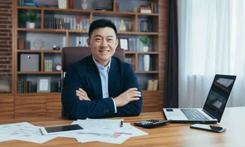 A tax professional smiling at the camera while seated at his office desk with an open laptop.